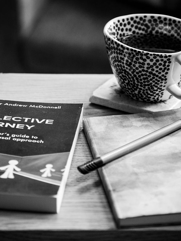 Black and white image showing a side table with a cup of coffee and a copy of The Reflective Journey by Professor Andrew McDonnell